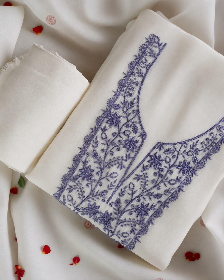 An off-white fabric garment or textile laid flat, featuring a prominent blue floral and vine embroidery design on the front panel. A matching rolled piece of the same fabric sits beside it on a draped white background with scattered red flower petals.